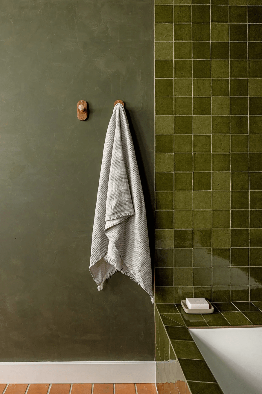 Towel hanging on a hook on a green wall next to green tiled wall, with a white object on a ledge.