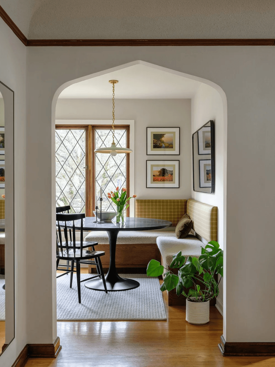 Living room with window, dining table, chairs, and framed pictures on wall, decorated with plants and a yellow cushioned bench.