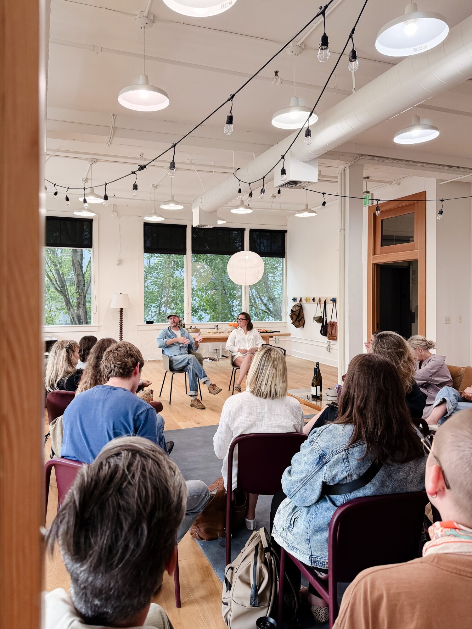 People seated in a room with large windows, listening to a speaker at the front, with hanging lights and a wooden door.