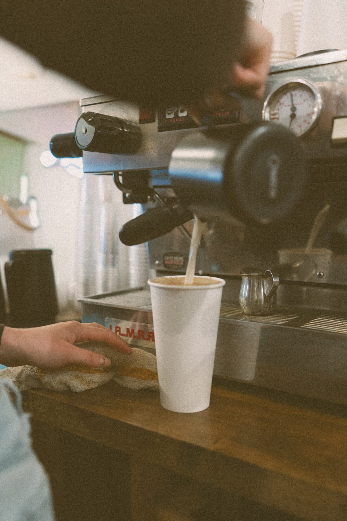 A barista prepares a coffee shot into a tall white cup on a wooden counter, with coffee equipment in the background.