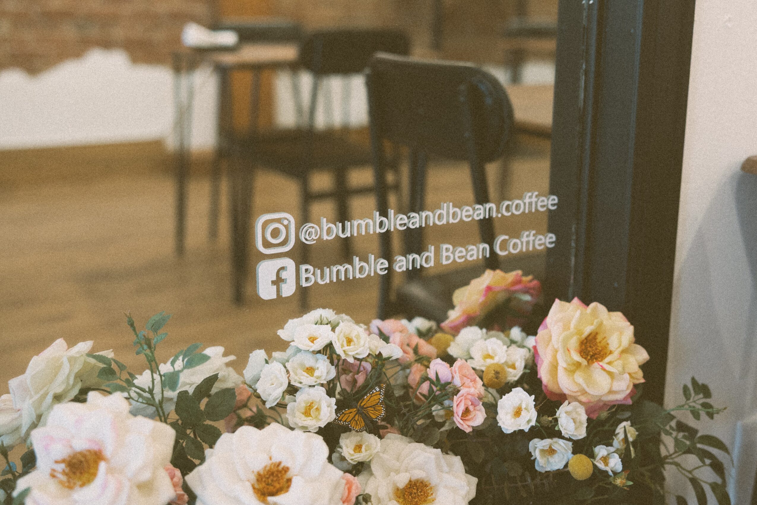 Close-up of a window with flowers outside, social media handles for Bumble and Bean Coffee, interior of a cafe visible through the window.