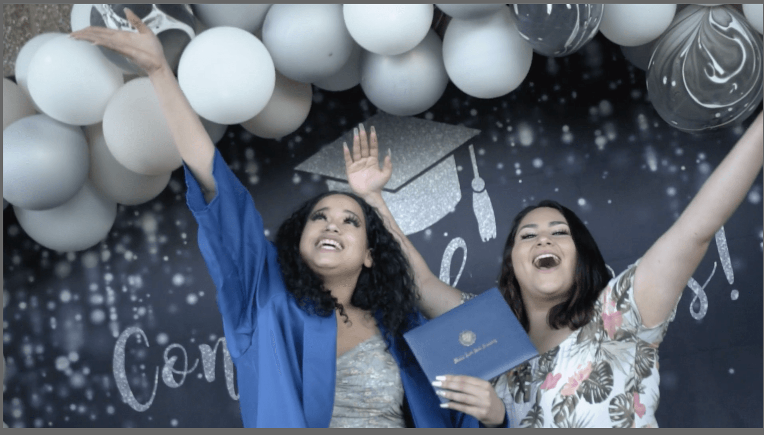 Two women celebrating with balloons and decorations, smiling and raising their hands, one holding a diploma or certificate.