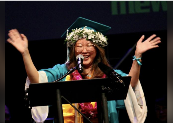 Person wearing a graduation cap and gown, smiling, with arms raised, standing behind a podium, decorated with flowers.