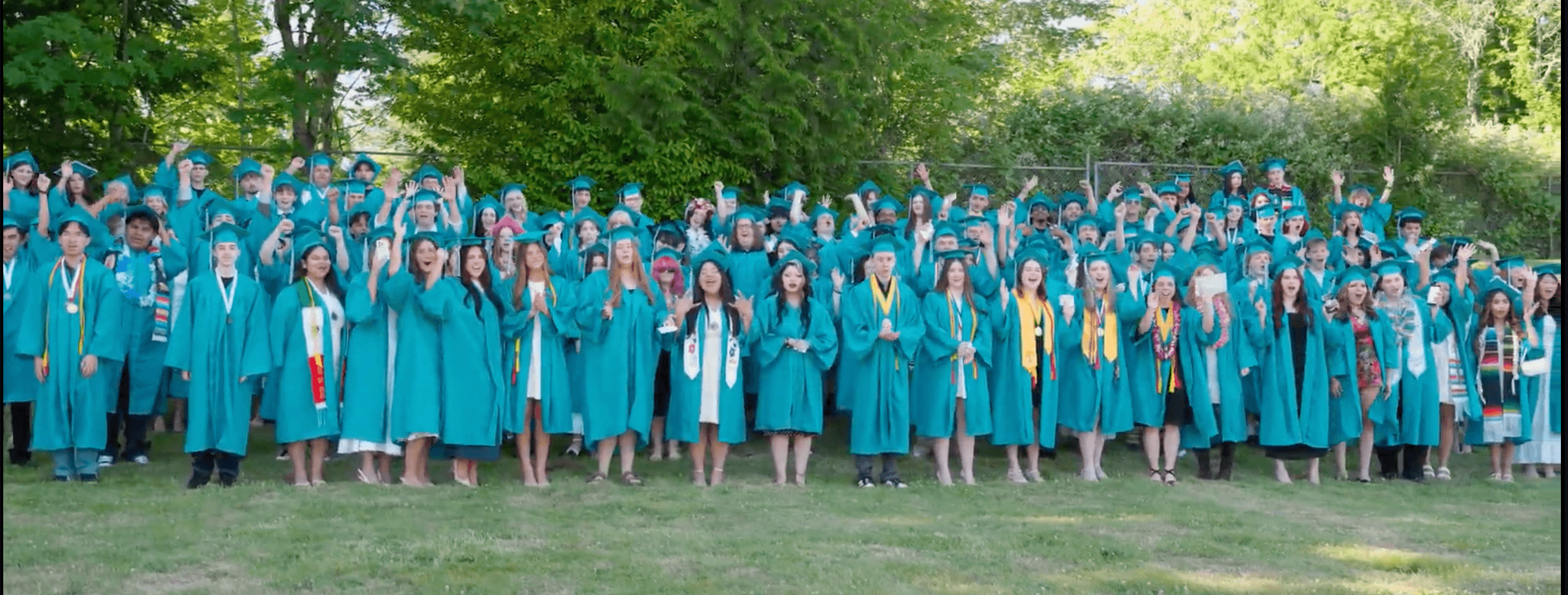 Group of graduates in blue caps and gowns outdoors, standing on grass with trees in background.
