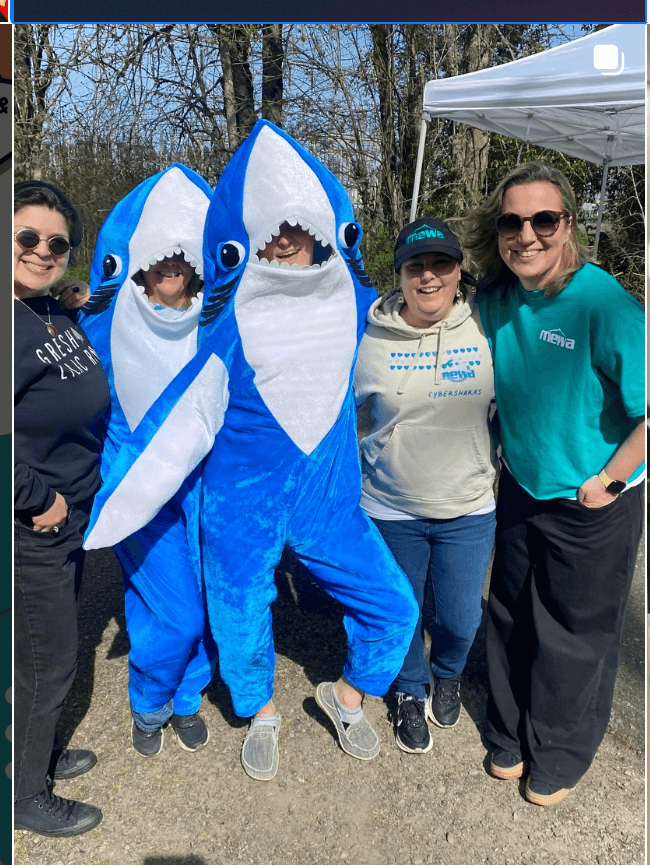 Three people standing outdoors with two people in shark costumes, smiling, with trees and a tent in the background.