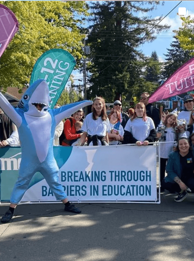 People gathered outdoors behind a banner with a shark mascot, trees, and tents in the background.