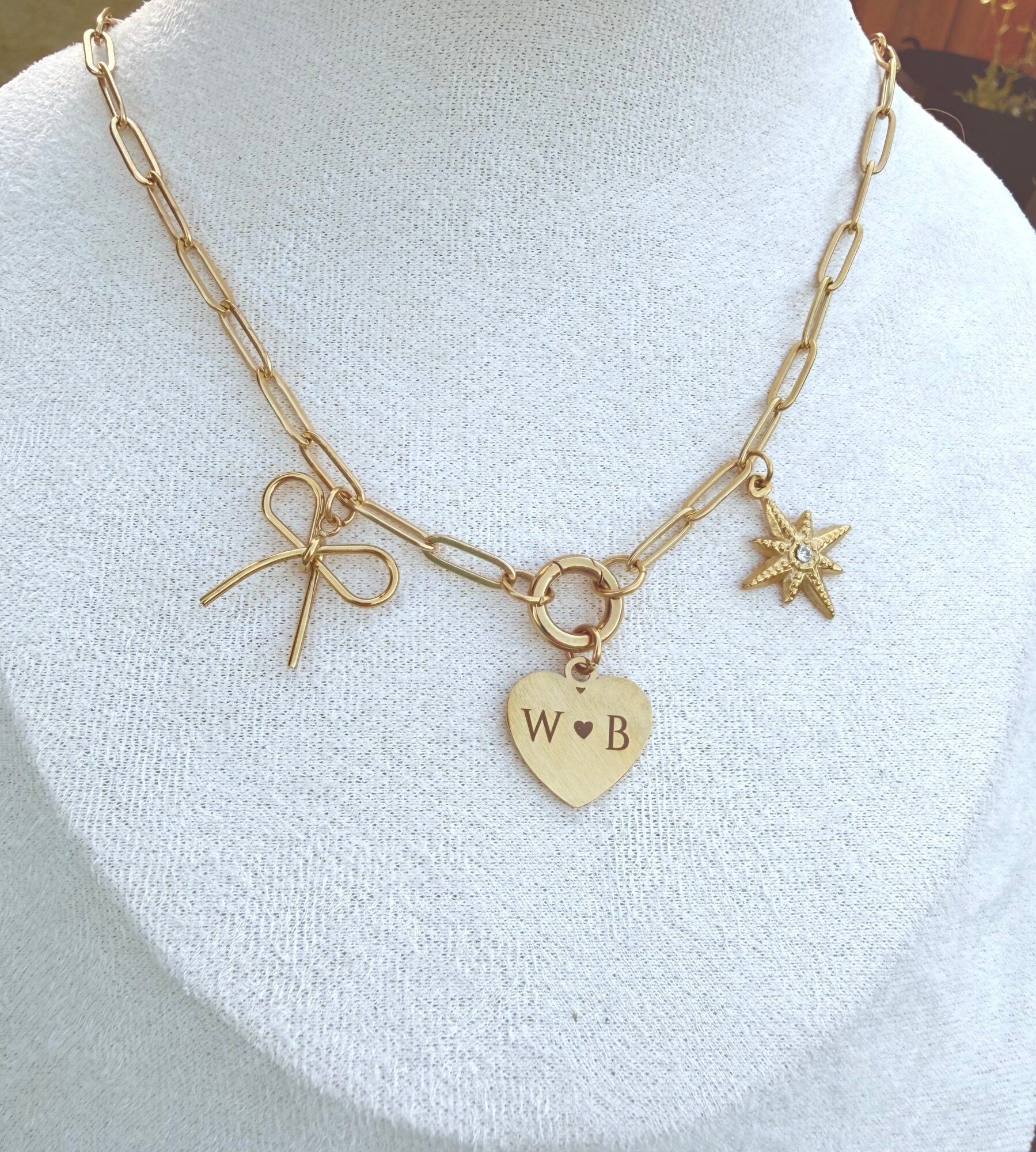 Gold chain necklace with a heart-shaped pendant engraved with 'W B', a star charm, and a bow charm, displayed on a white bust.