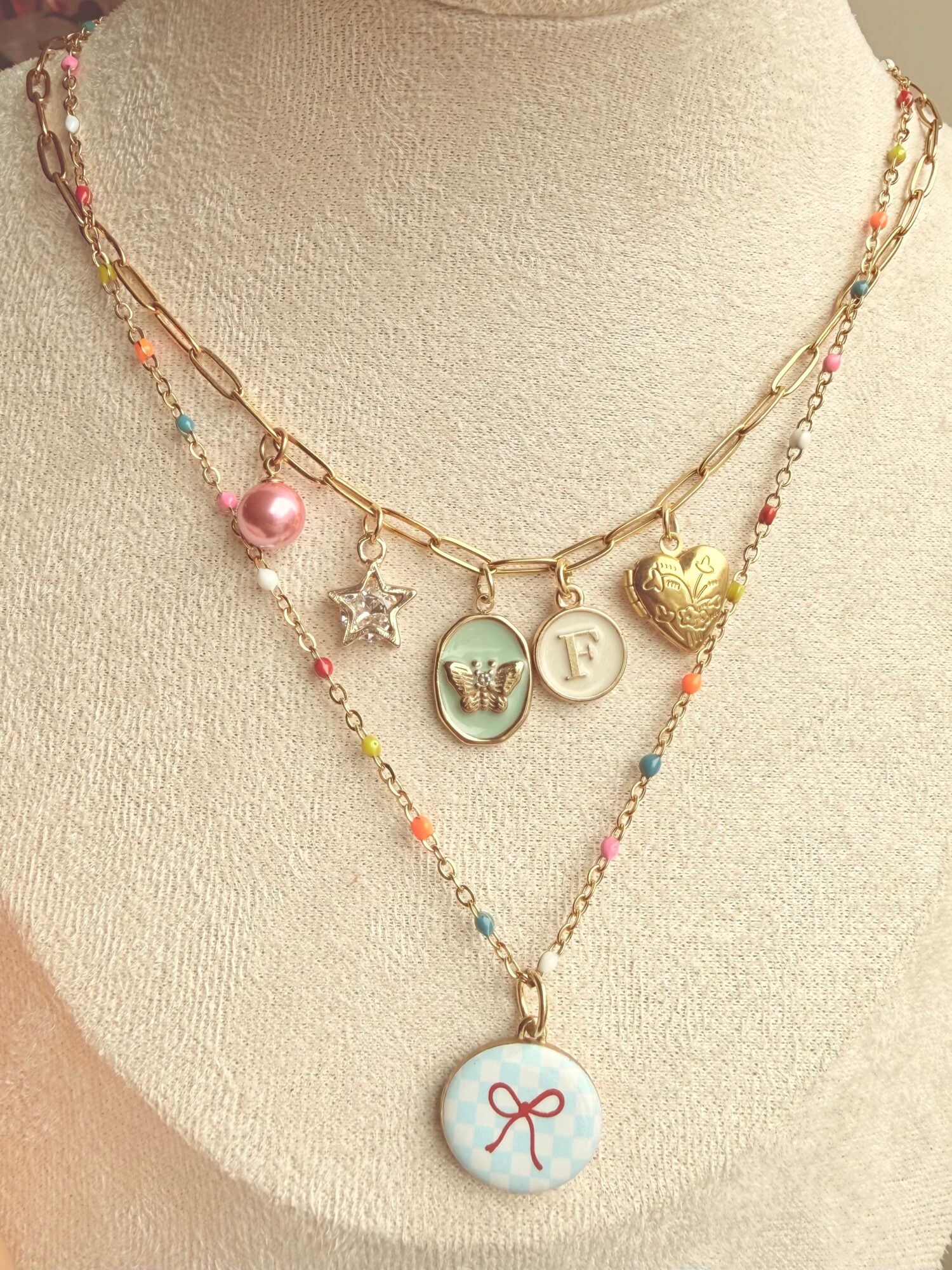 Two layered necklaces with colorful beads and charms, including a pink pearl, star, heart, and circular pendant with a bow, on a beige bust.