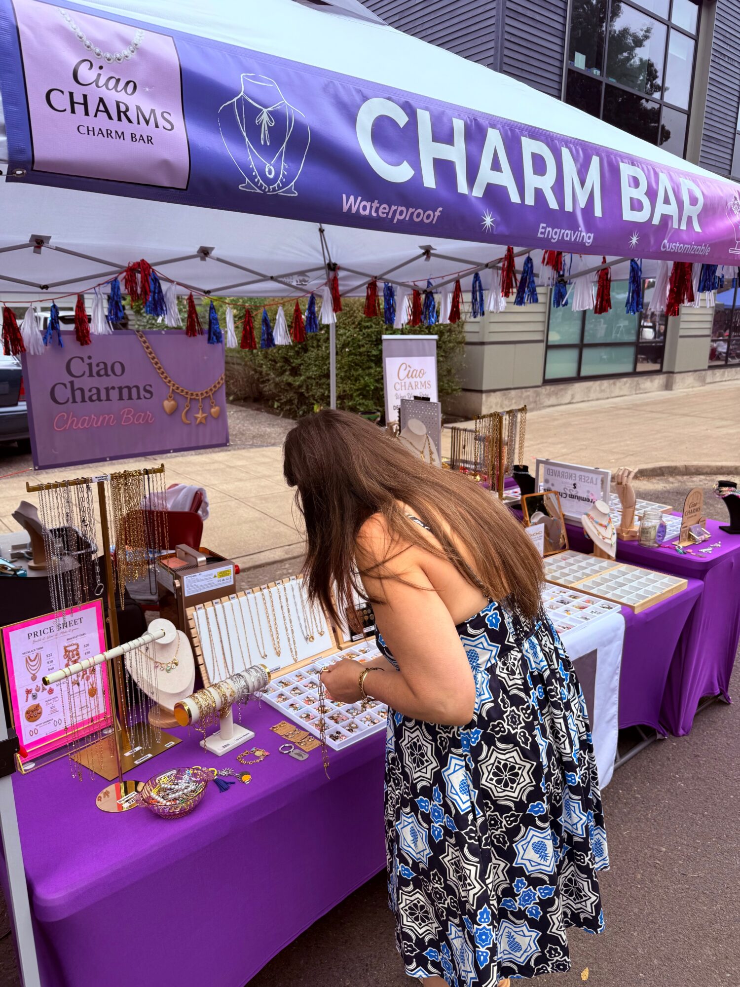 Woman in a patterned dress browsing jewelry display at outdoor Charm Bar booth with purple tablecloths and banners.