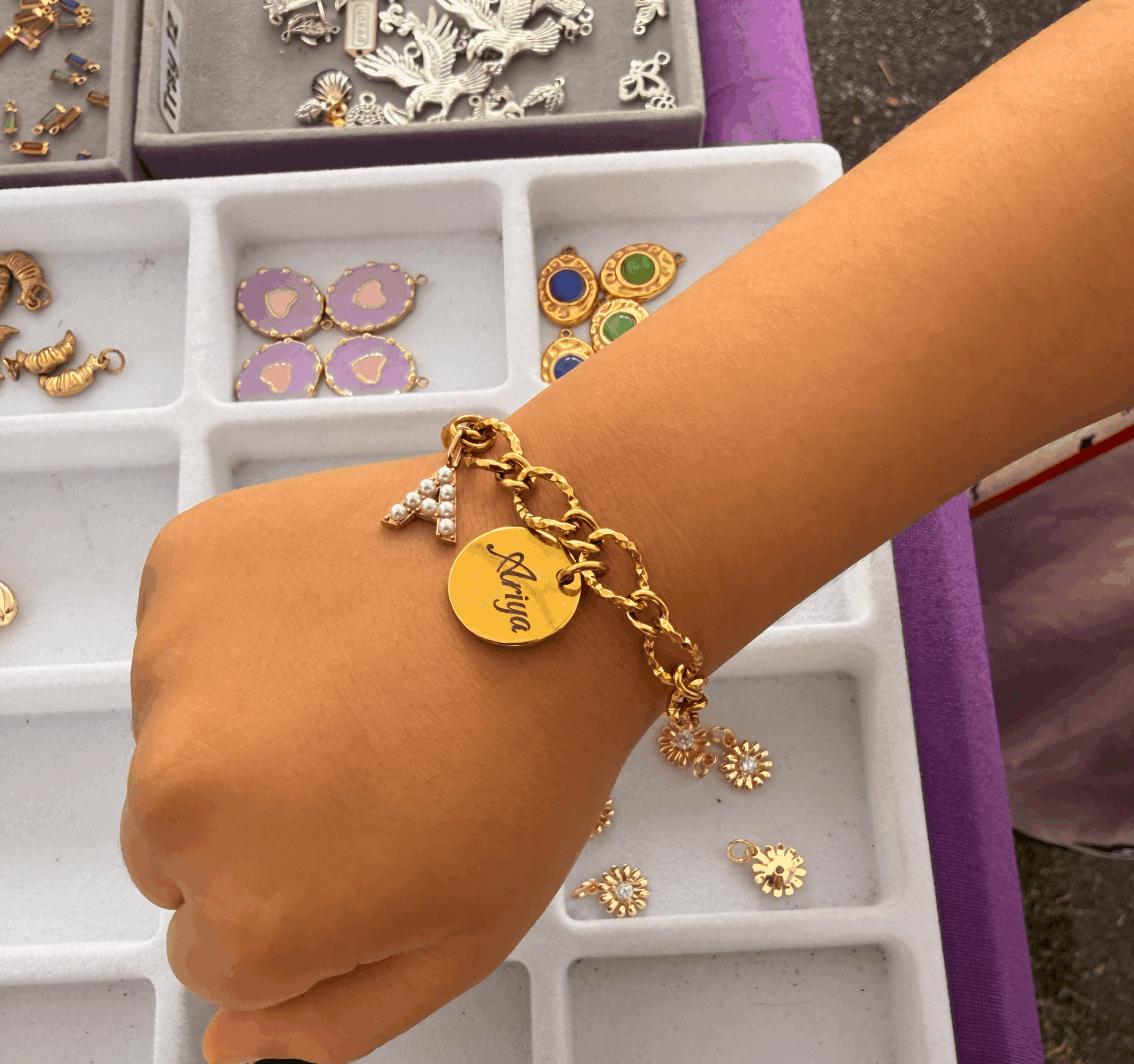 Person's wrist with gold bracelet and charm, jewelry tray with earrings and charms in background.