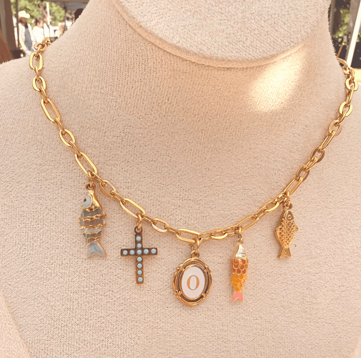 Gold chain necklace with five charms, including a fish, cross, letter O, and two other fish, displayed on a beige mannequin.
