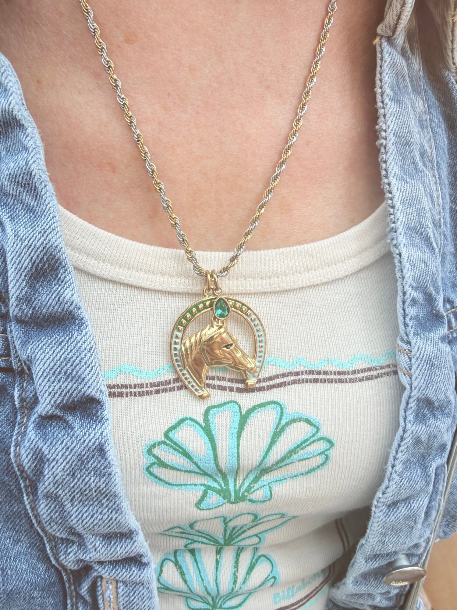 Close-up of a person wearing a gold necklace with a circular pendant and a turquoise stone, over a white shirt with a floral design, and a denim jacket.