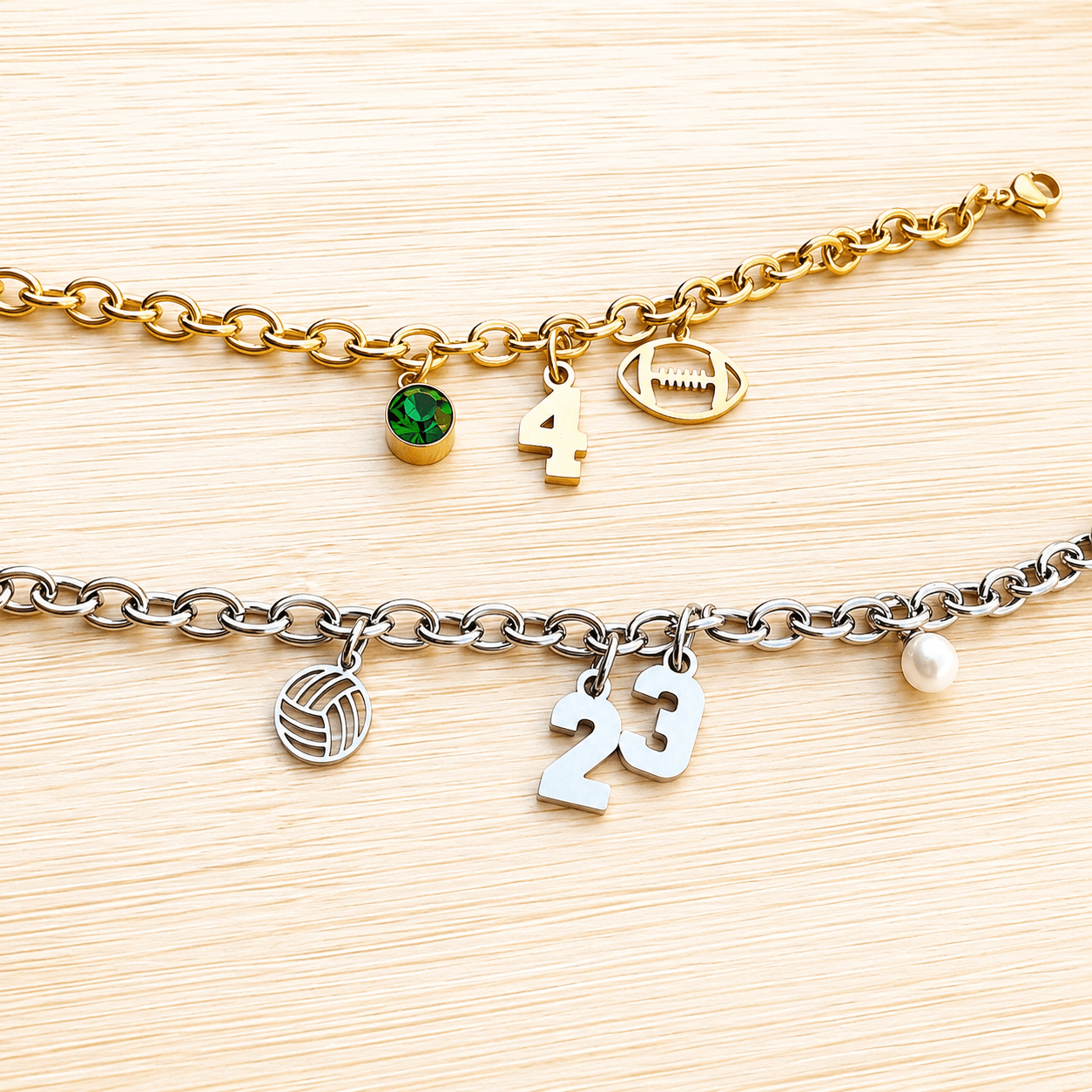 Two bracelets with charms and numbers on a light wooden surface, one gold with a green gem, the other silver with a pearl.