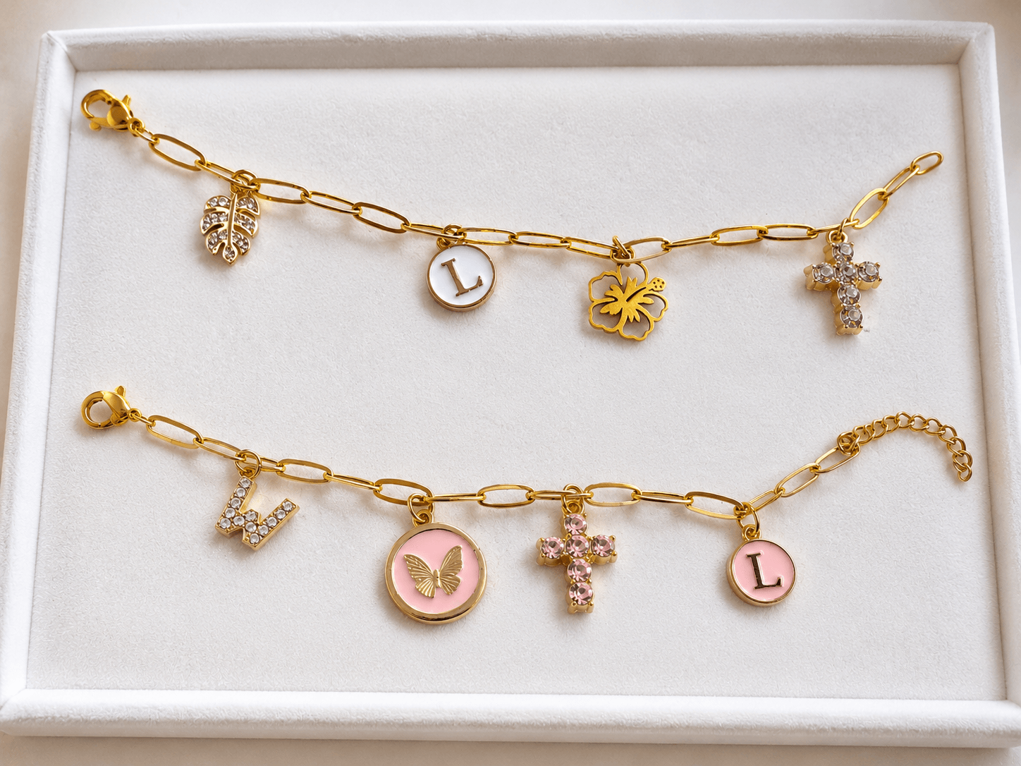 Two gold charm bracelets with various charms, including a butterfly, cross, and letter L, displayed in a white box.