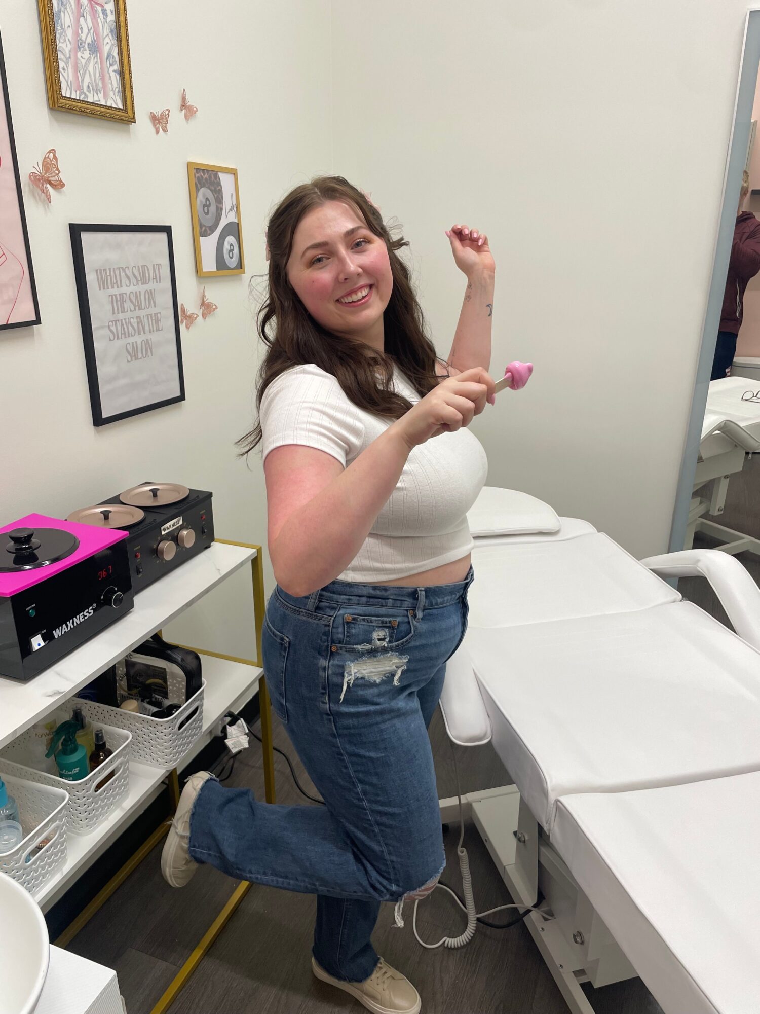 Young woman with long brown hair smiling, holding a pink object, standing next to a hospital bed in a medical room.