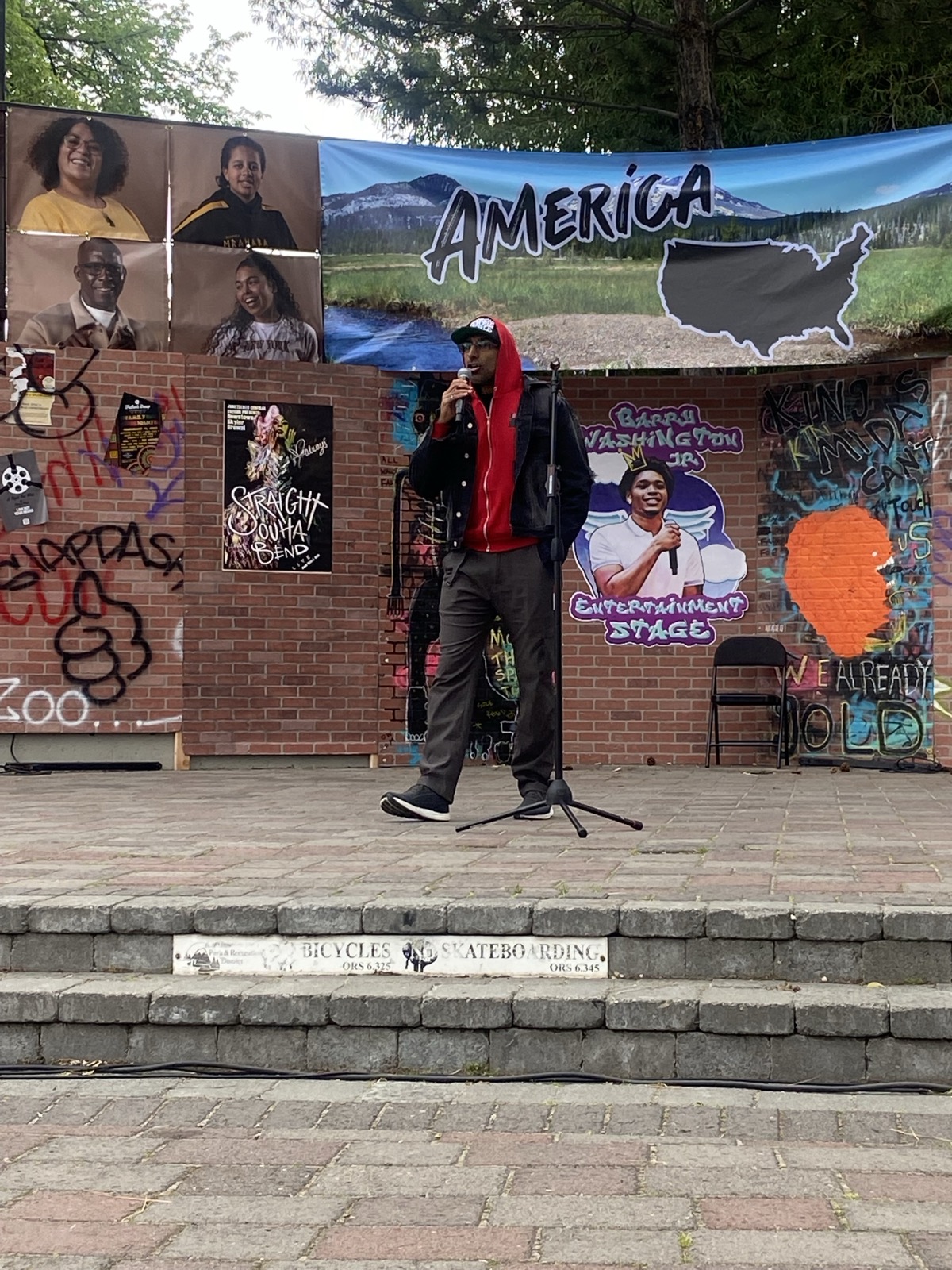 Person speaking on stage with a microphone, backdrop features images of people and a map of the United States, outdoors setting.