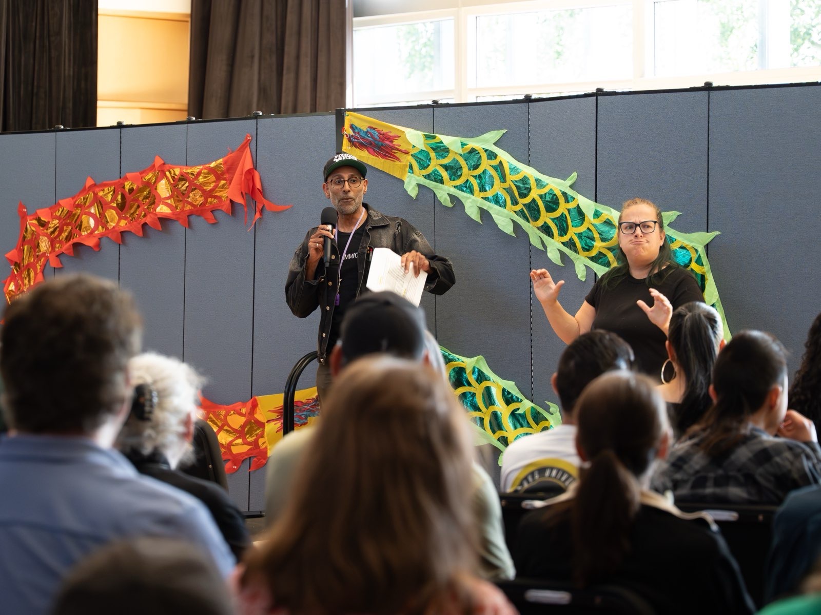 Two people stand on stage with colorful dragon decorations, speaking to an audience in a room with large windows.