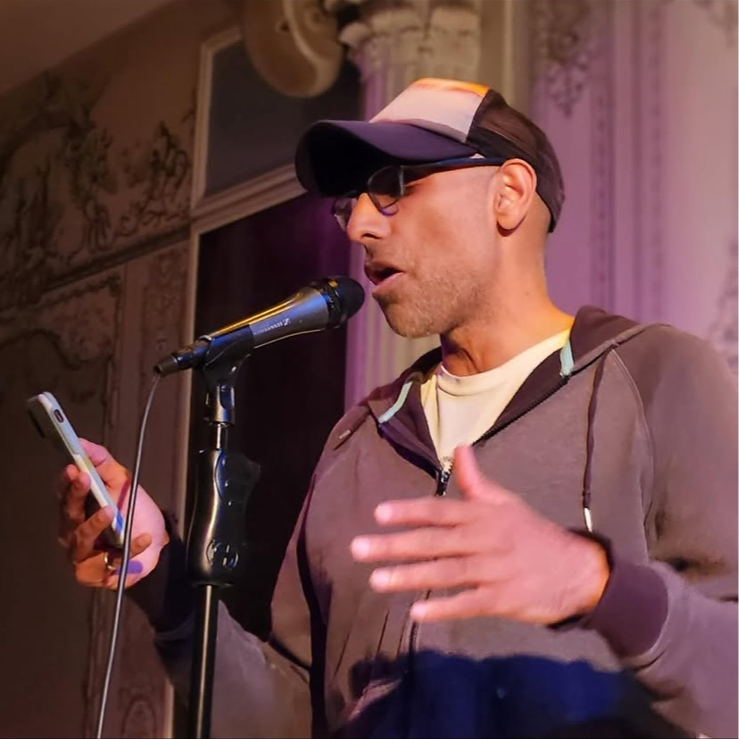 Man wearing glasses, cap, and hoodie speaking into microphone, holding a phone, gesturing with his hand.