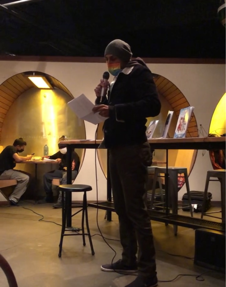 Person wearing a mask and beanie speaking into a microphone, holding papers, in a dimly lit room with arched wall features.