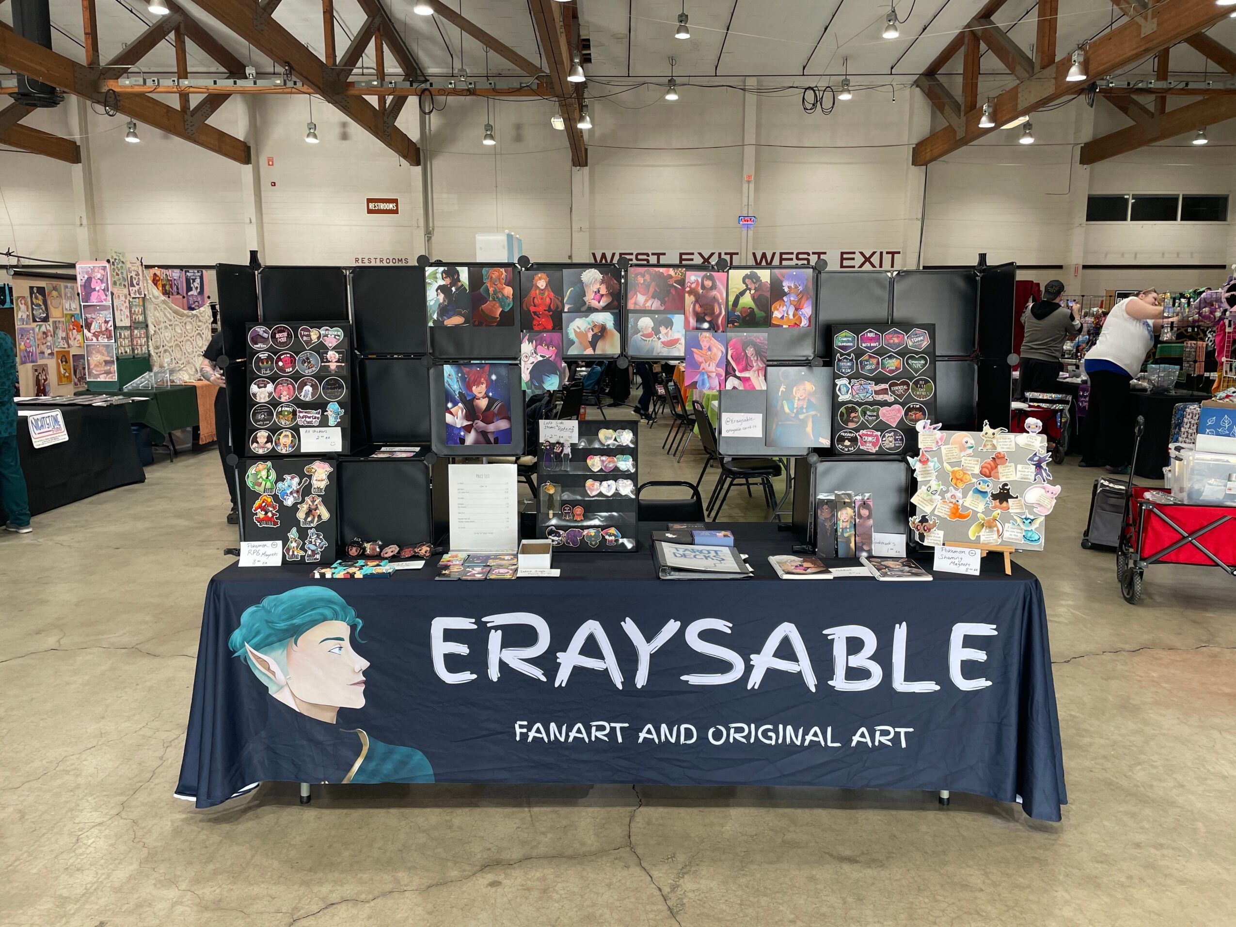 Vendor booth with artwork and merchandise, table with a blue cloth displaying 'ERAYSABLE' and a character illustration, in an indoor event space.