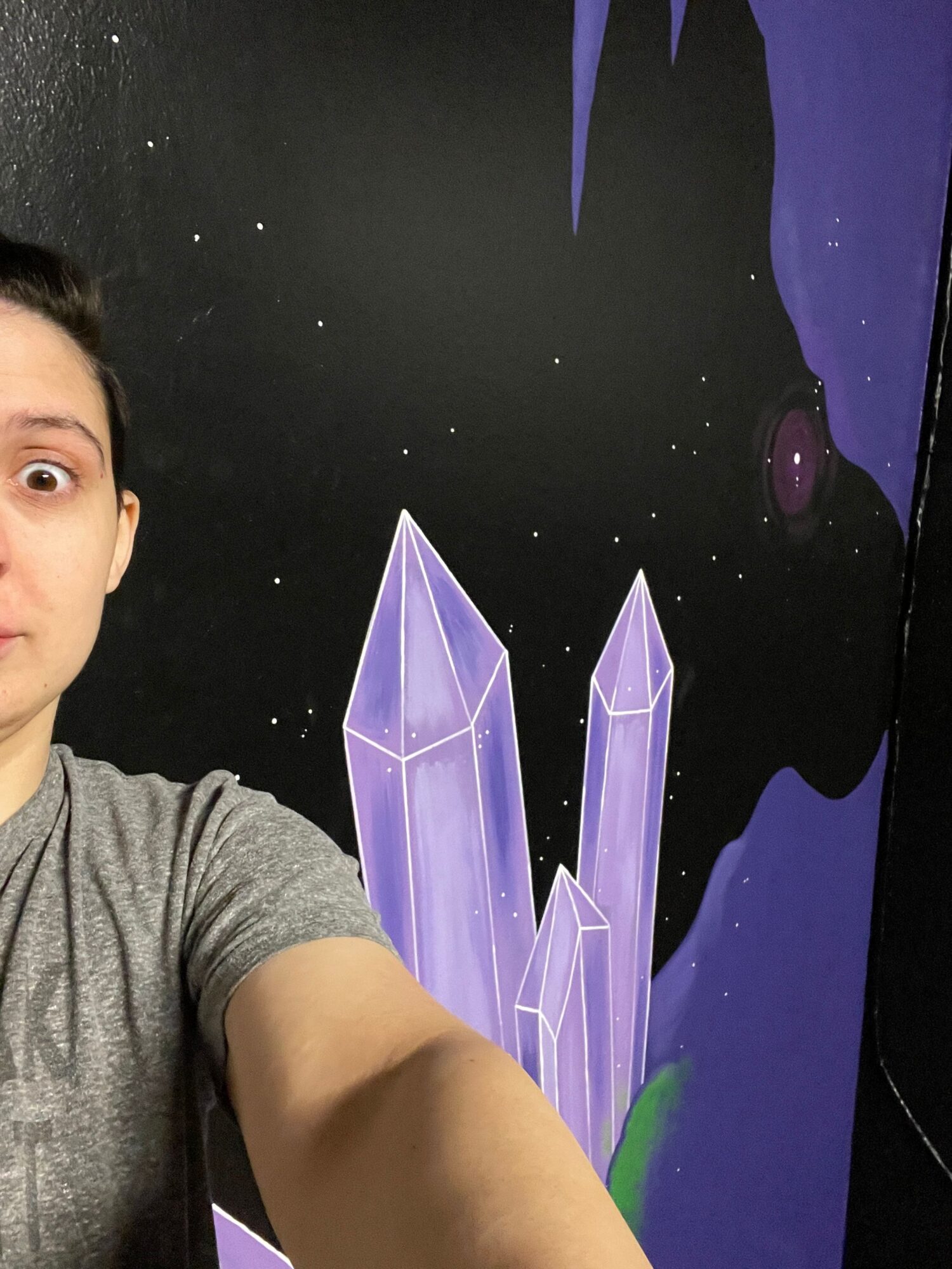 Part of a person’s arm and shoulder in a gray shirt, with a space-themed mural featuring purple crystals and stars in the background.