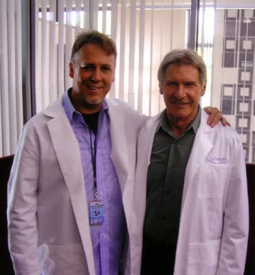 Two men in white lab coats standing indoors, smiling, with vertical blinds behind them.