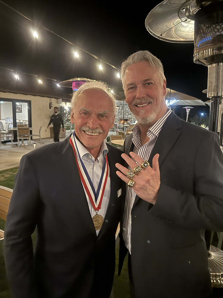Two men in suits smiling at an outdoor event at night, with string lights overhead and a large outdoor heater nearby.