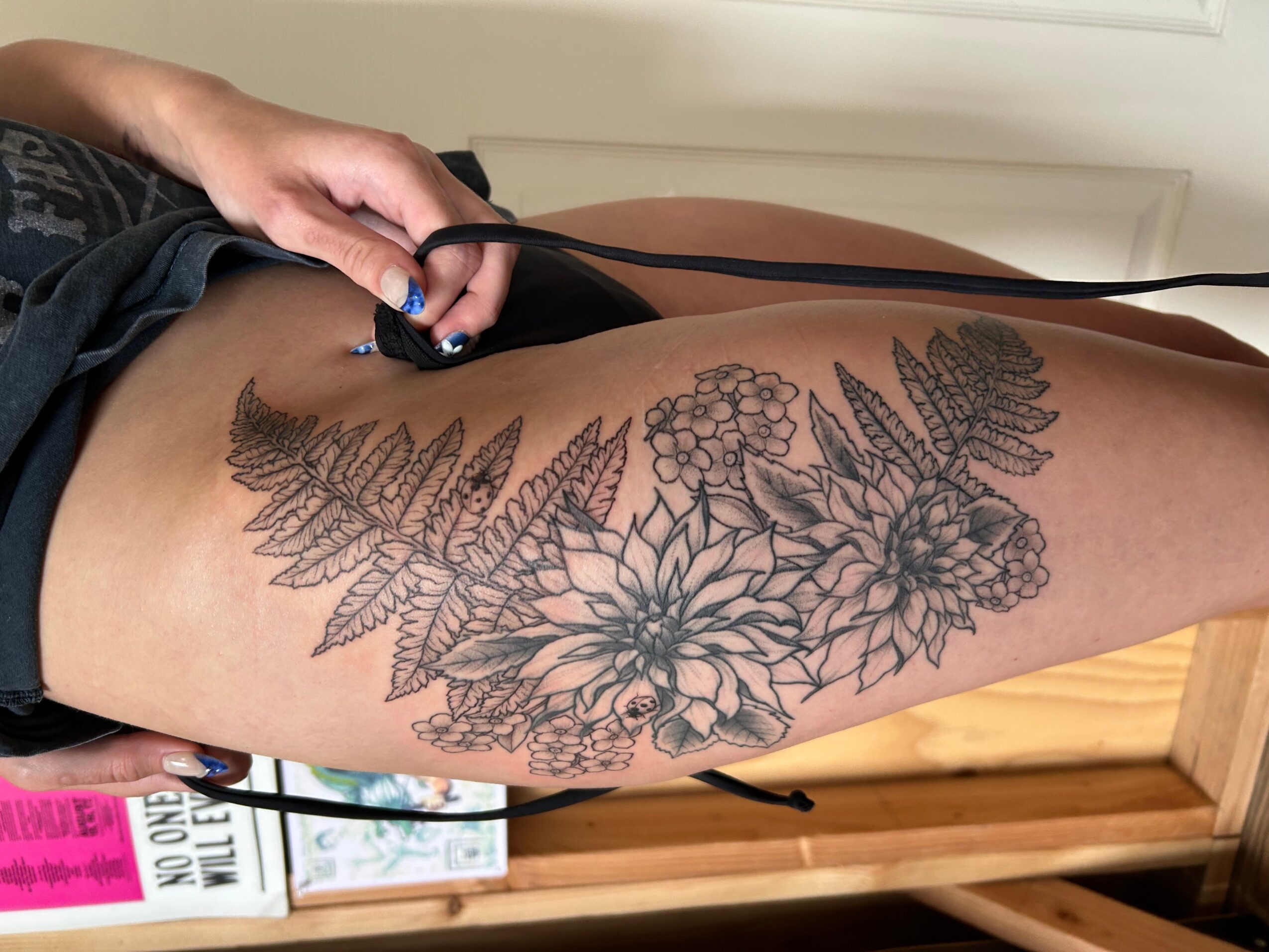 Person lying down with a floral tattoo on thigh, holding a device, background includes a wooden surface and books.