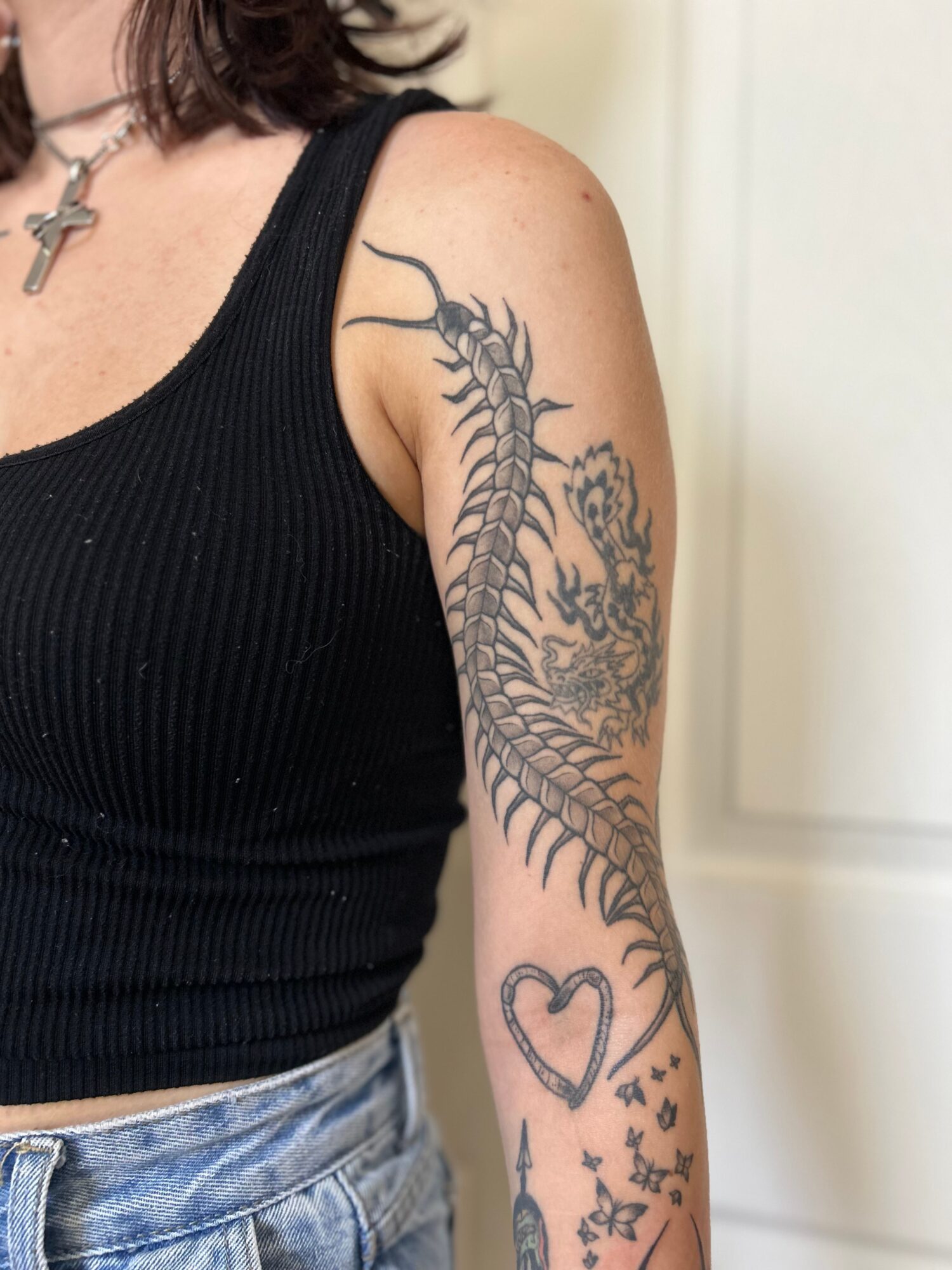 Person with a tattoo of a dragon on their upper arm and a heart with stars on their forearm, wearing a black sleeveless top.