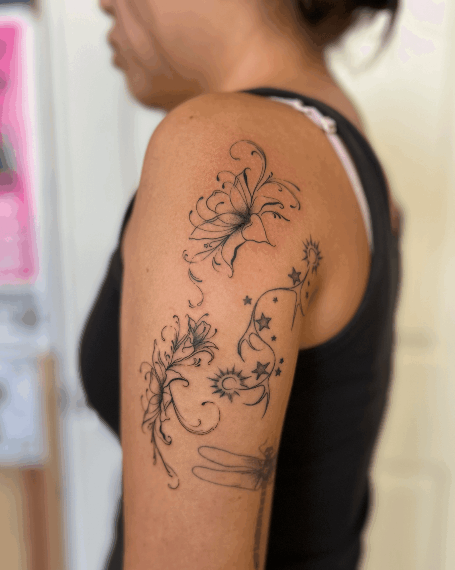 Tattoo of a butterfly, flowers, and stars on person's upper arm, with a black sleeveless top, background blurred.