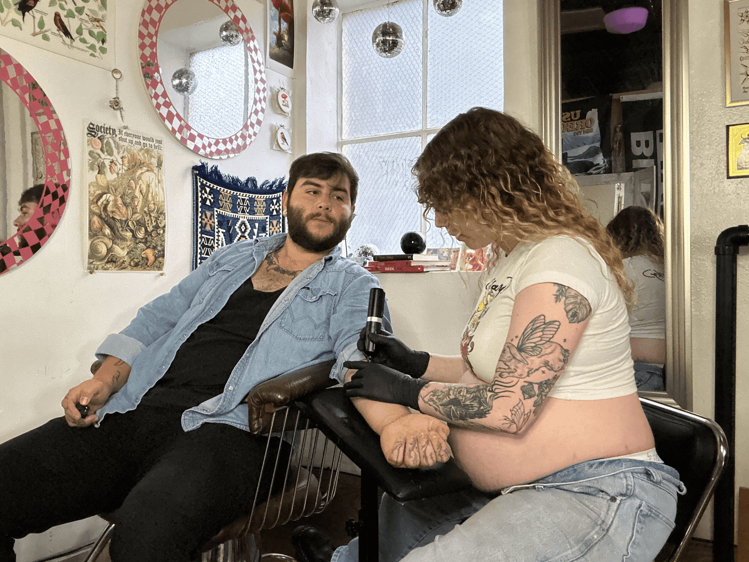 A person with tattoos on their arm is getting a tattoo from a tattoo artist, who is looking at the client. They are in a tattoo studio with decorated walls and a window.