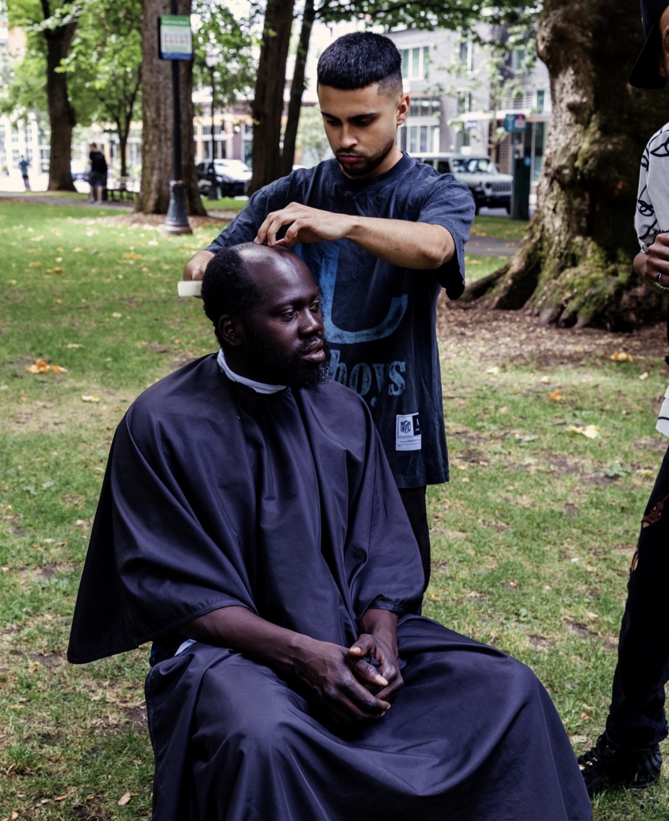 Man receiving a haircut outdoors in a park with trees and grass, surrounded by people. Short-haired barber cutting hair.