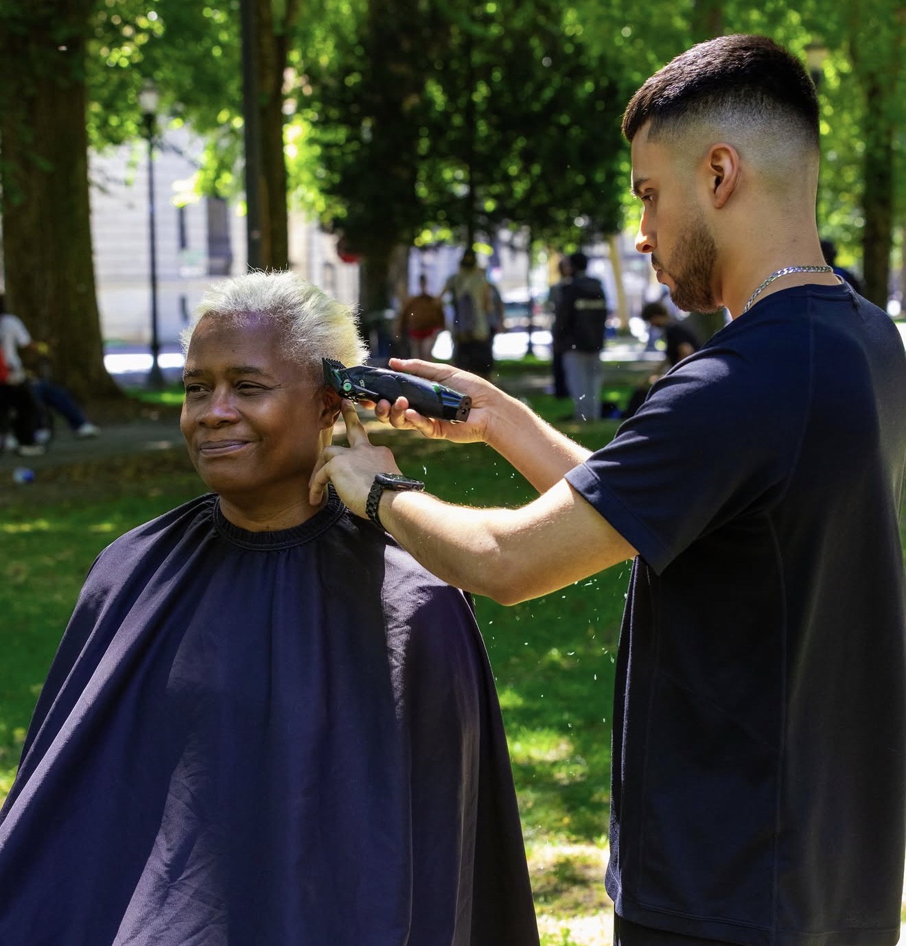 Young man giving a haircut to an elderly woman outdoors in a park with trees and people in the background.