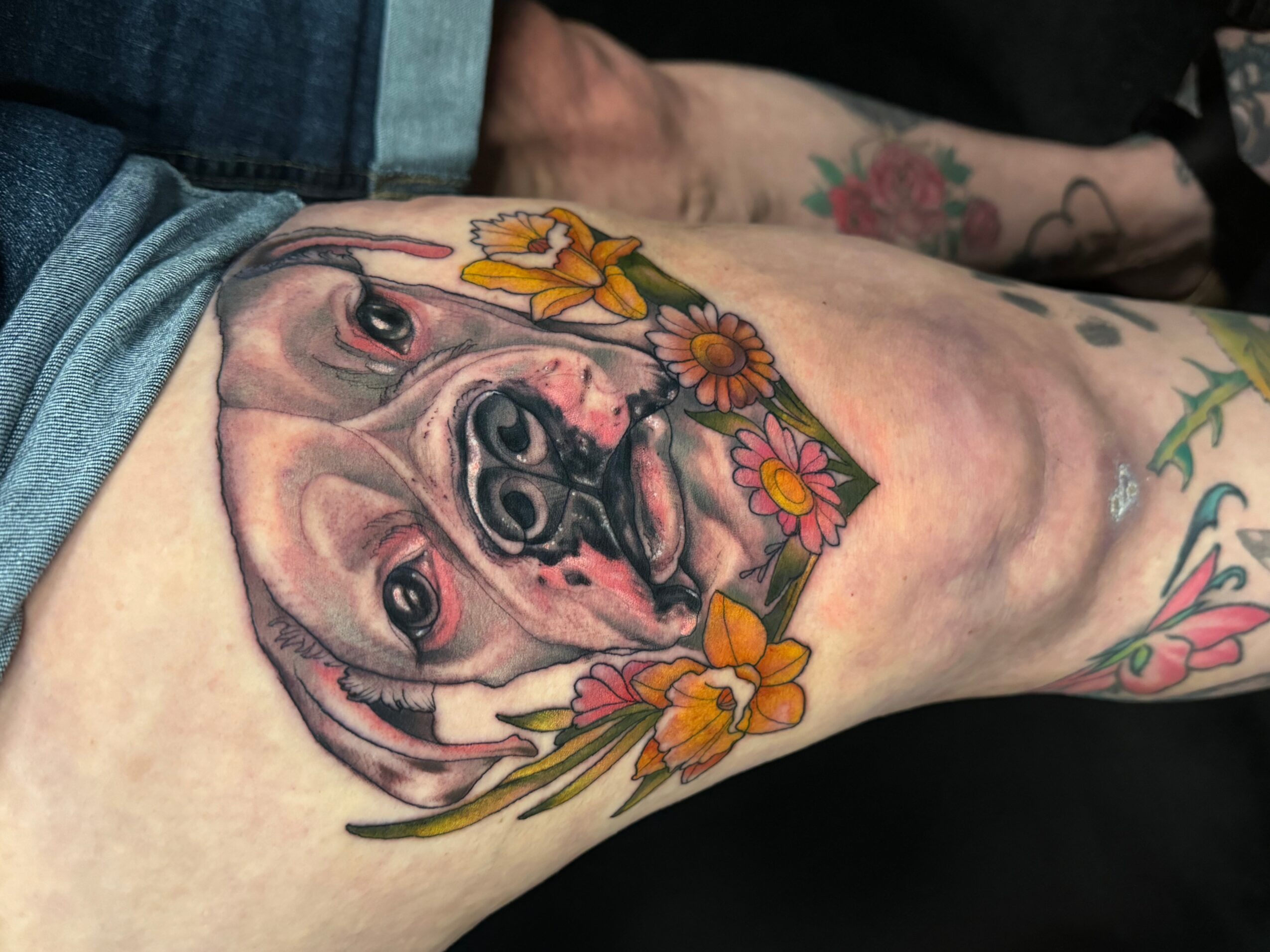Tattoo of a dog's face surrounded by colorful flowers on person's thigh.