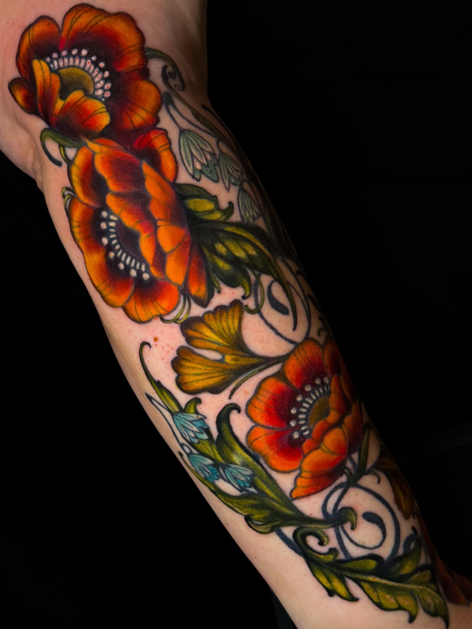 Tattooed arm with large orange and yellow flowers, green leaves, and black outlines against a black background.