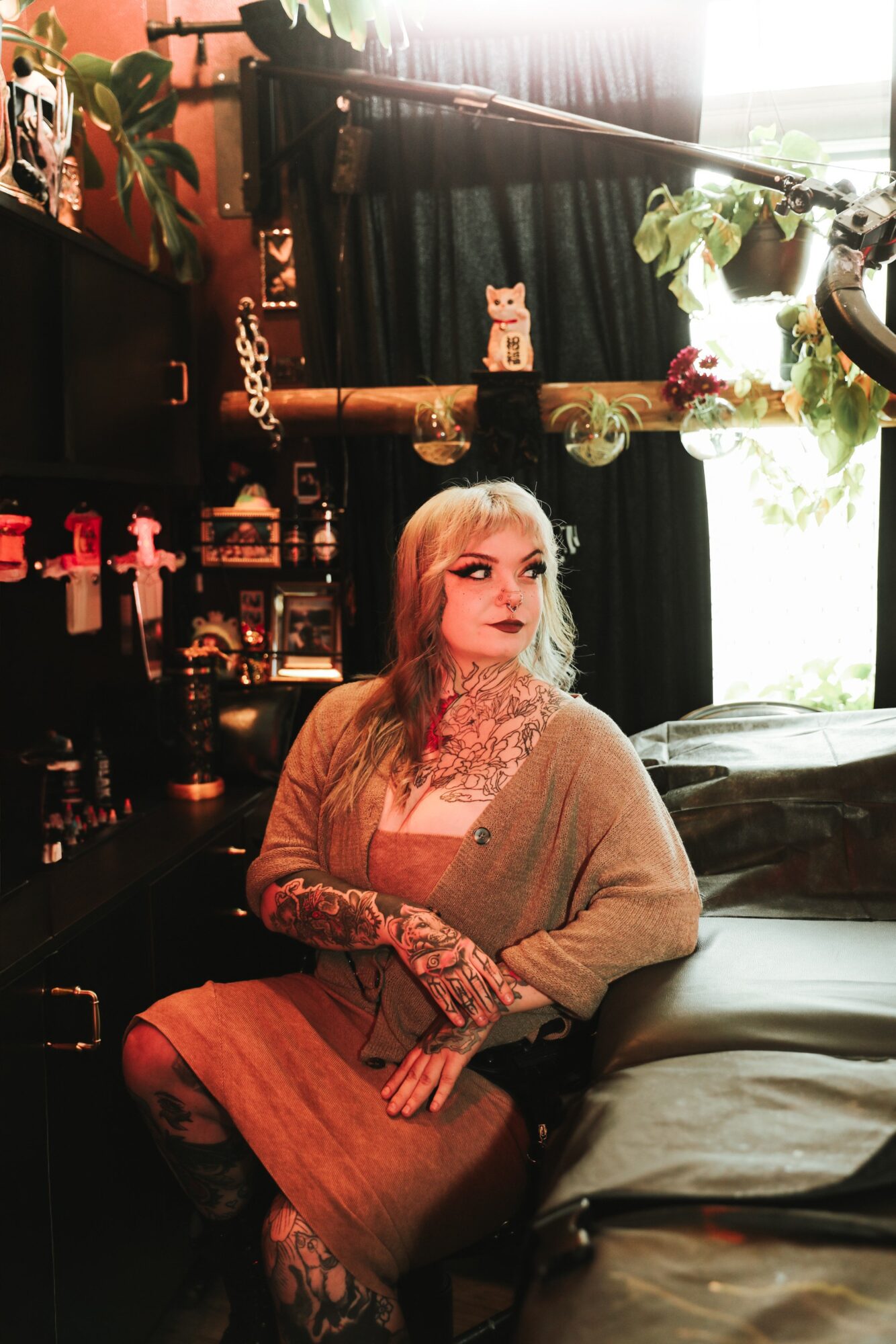 Woman with blonde hair and tattoos sitting on bed in a room with plants and decorative items, looking to the side.