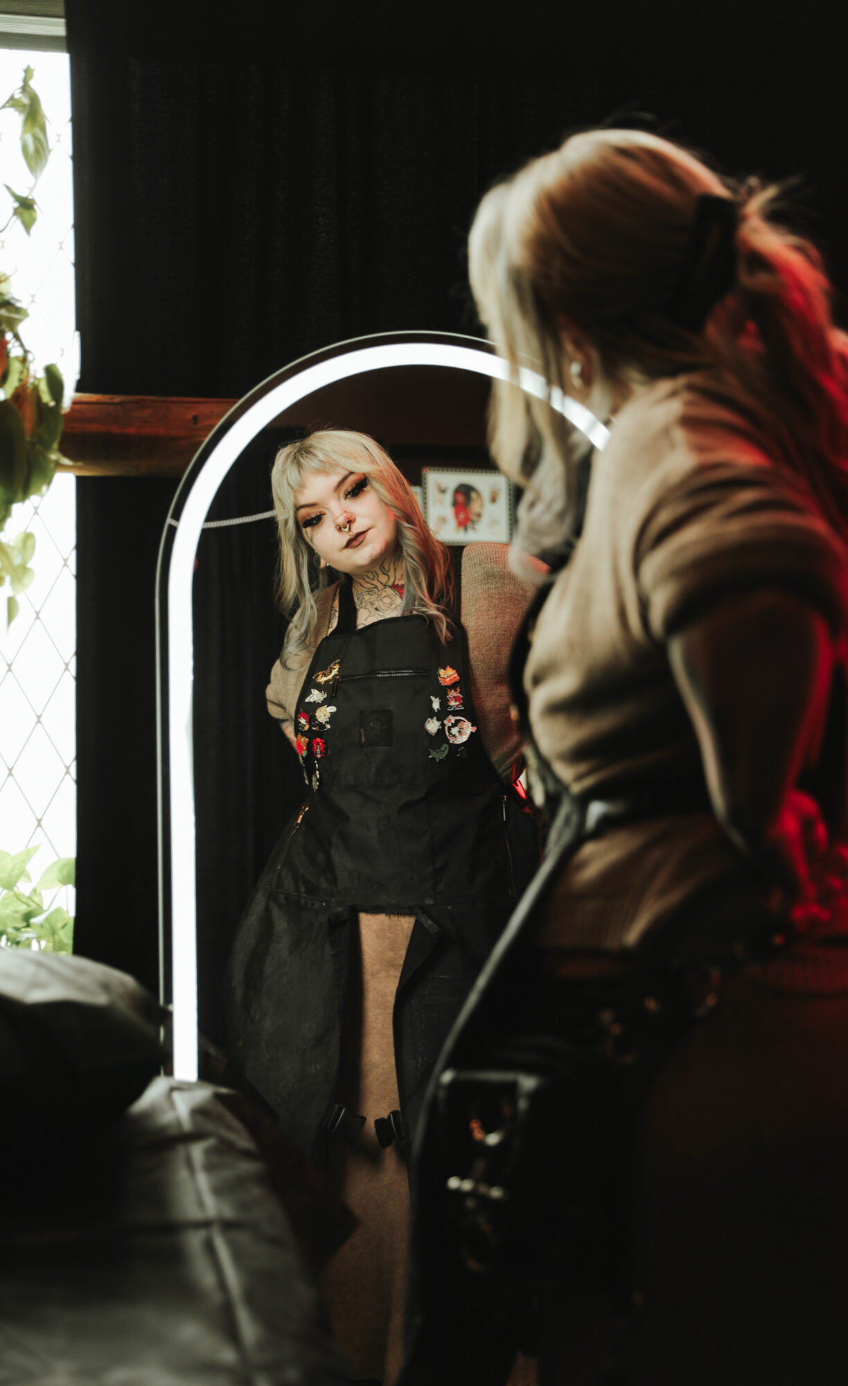 Person with brown hair in a ponytail looking at a mirror reflecting a woman with long blonde hair and dark clothing, in a room with dark curtains.
