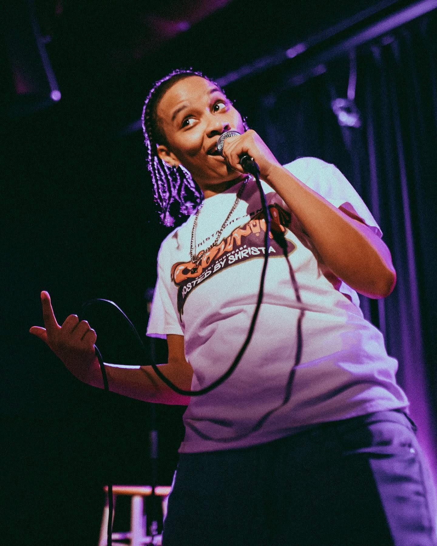 Person with purple braided hair holding a microphone on stage, wearing a white T-shirt and dark pants.