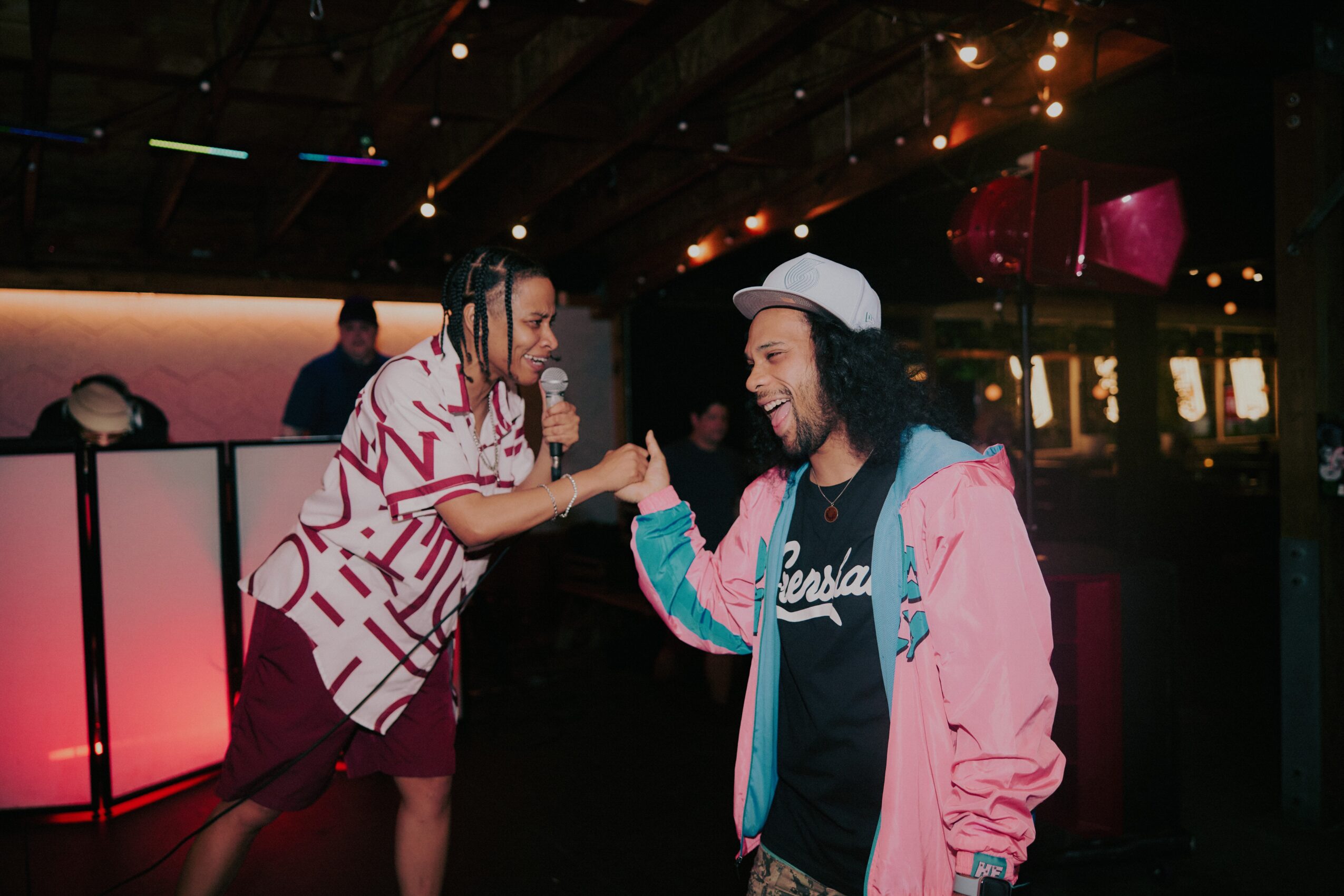 Two people dancing and smiling in a dimly lit indoor space with colorful lights, one wearing a patterned shirt and the other in a pink jacket.