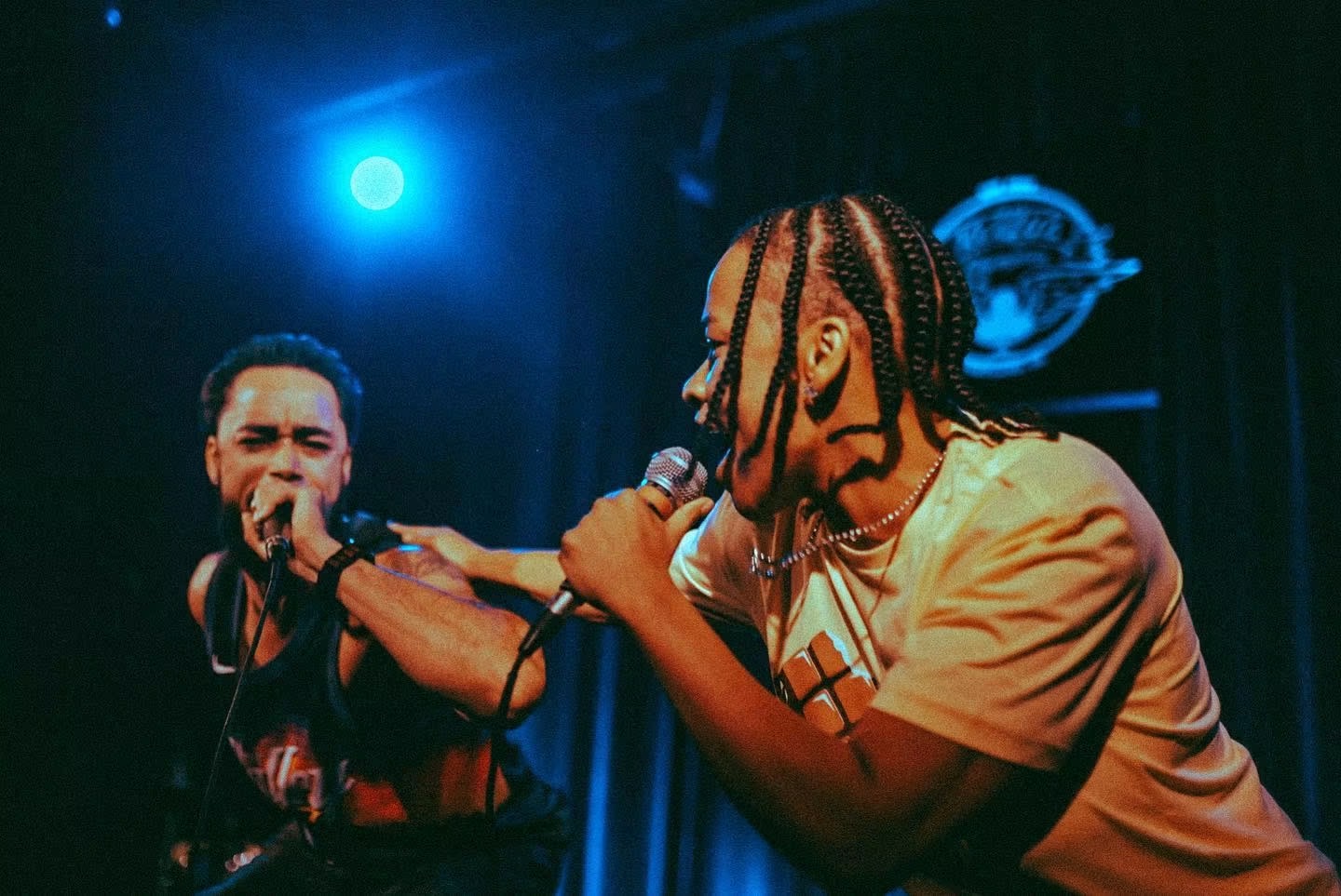 Two performers on stage, one singing into a microphone, with stage lighting and a logo in the background.