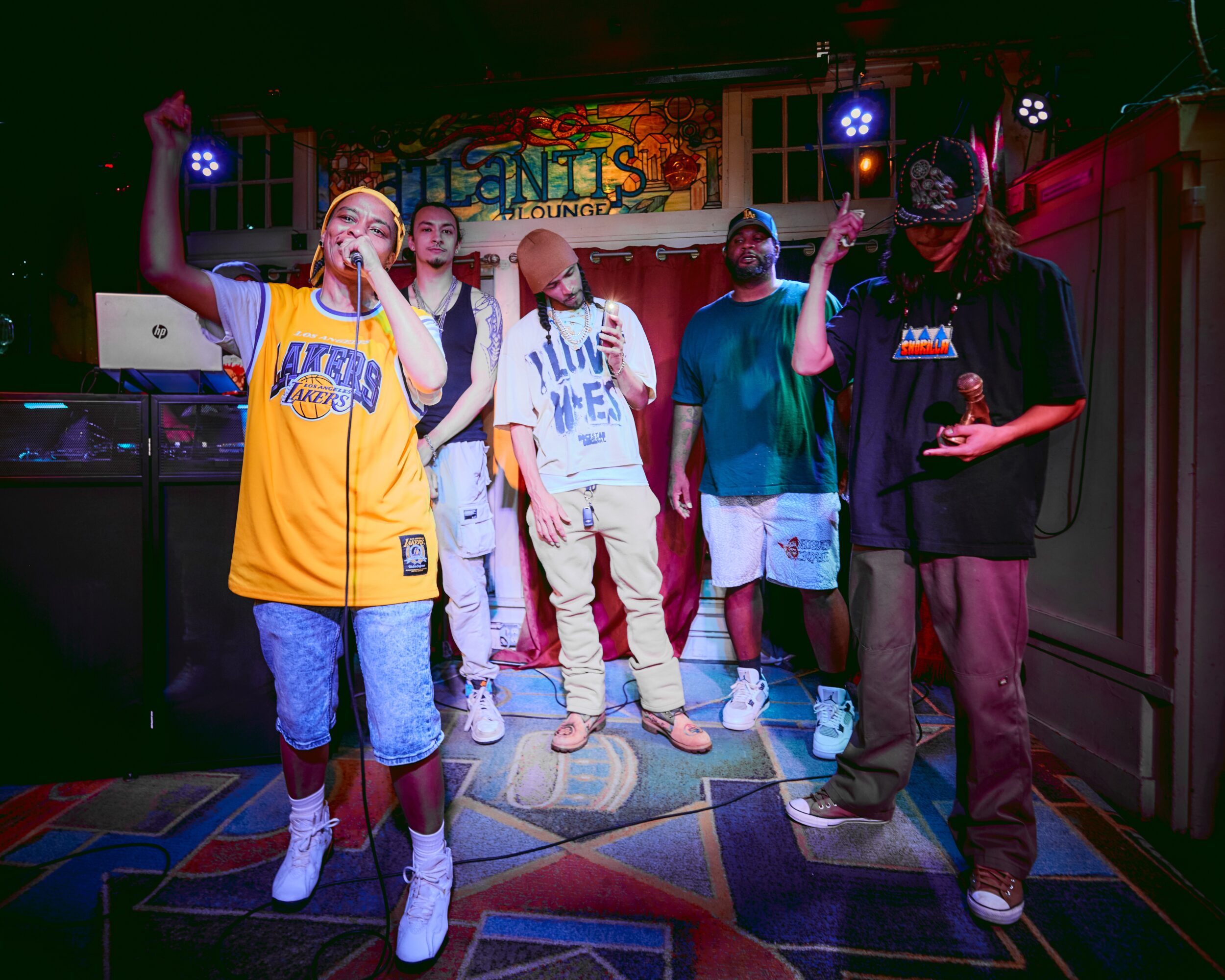 Group of five people performing on a stage with colorful lighting and a sign in the background, some raising hands.