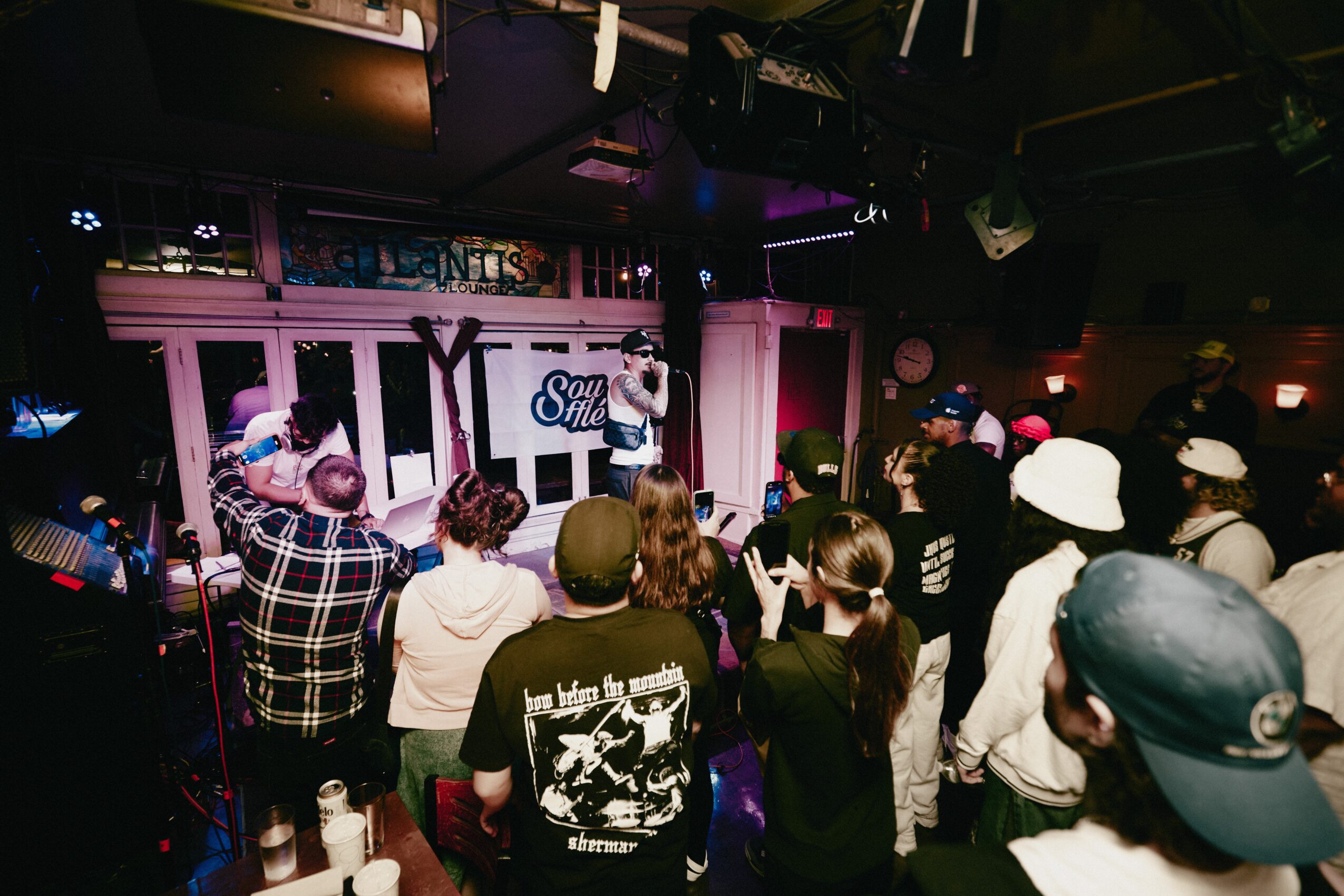 Person on stage speaking to an audience in a dimly lit venue with pink and purple lighting, audience watching attentively.