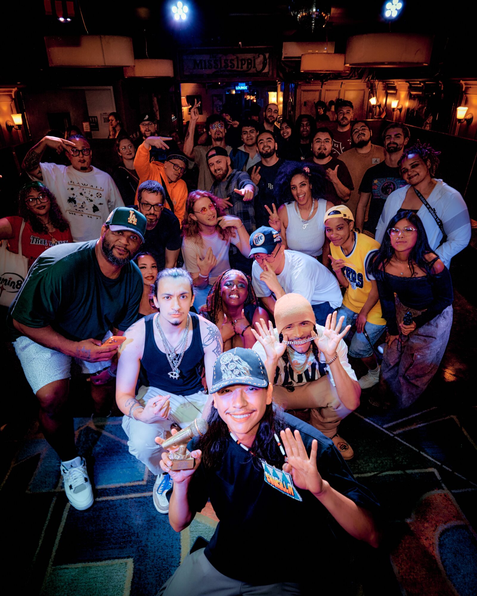 Group of diverse people gathered in a dimly lit indoor space, some smiling and making gestures, with a stage or performance area in the background.