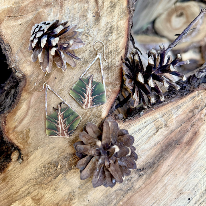 Pair of earrings with green and pink leaf design, pinecones, and a wooden background.