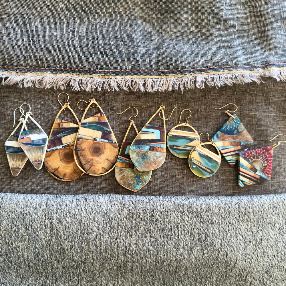 Seven pairs of colorful, patterned earrings with various shapes, laid out on a fabric surface.