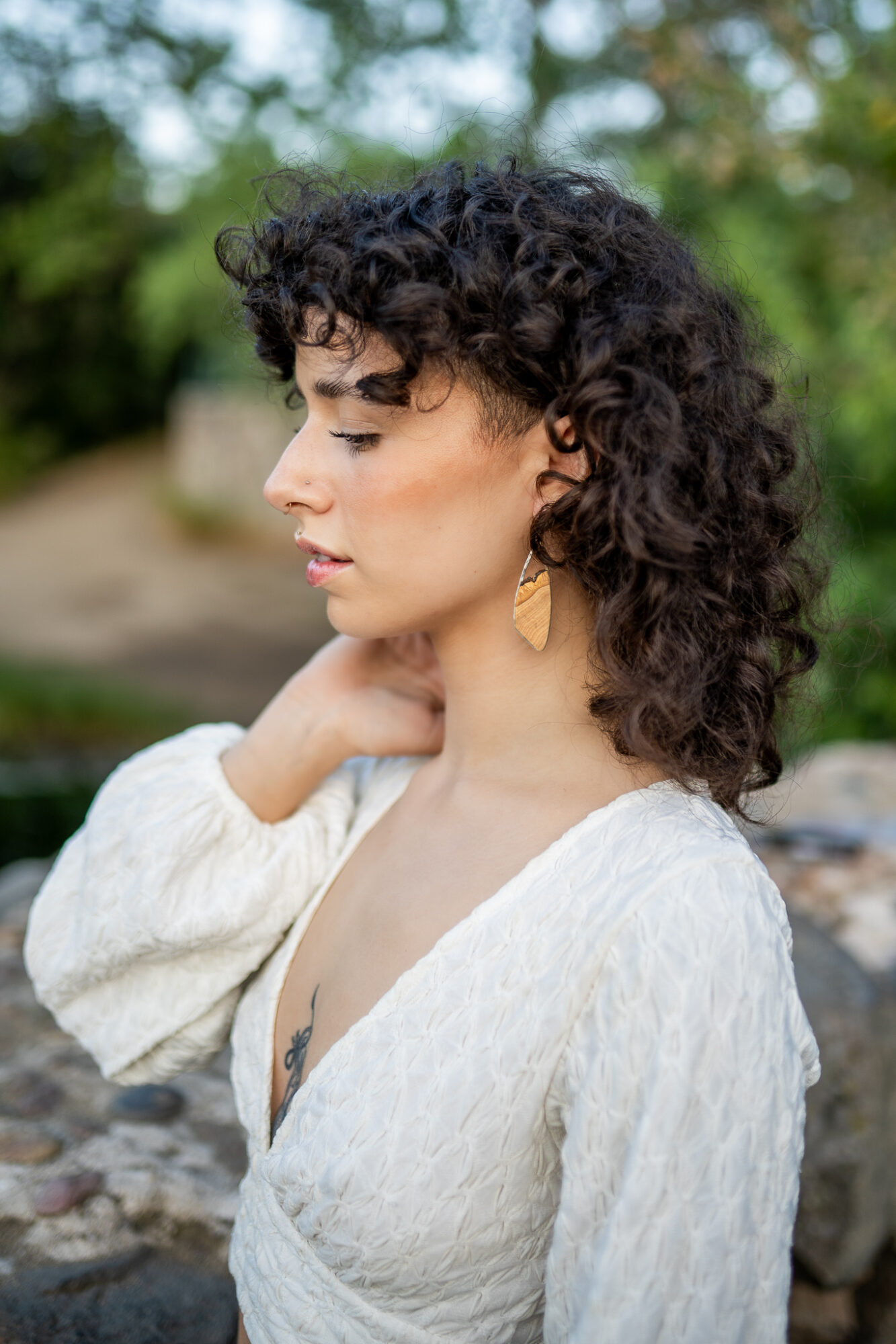 Profile of a woman with curly hair wearing a white top and gold earrings outdoors.