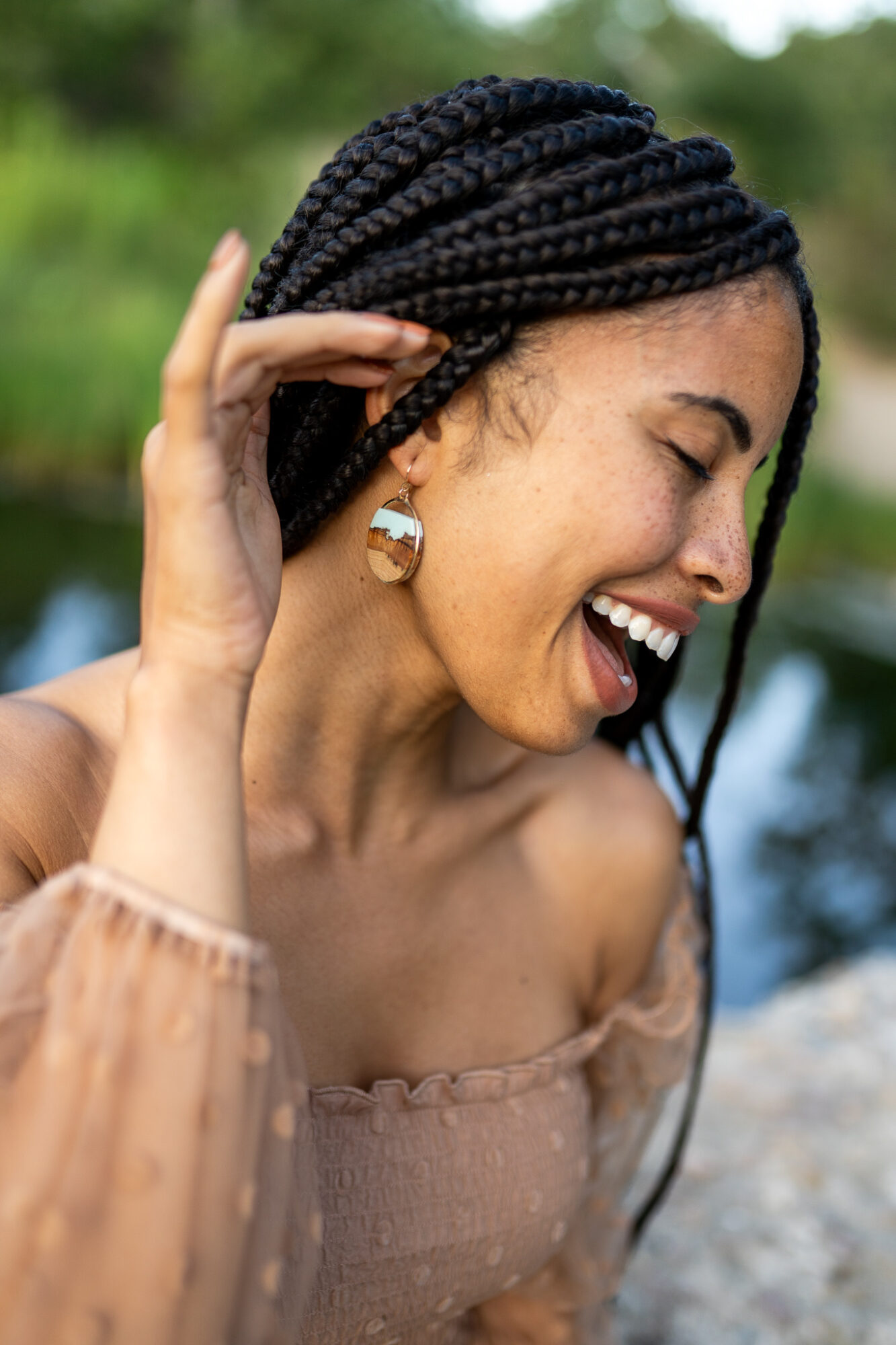 Smiling woman with braided hair outdoors, wearing earrings and a beige off-shoulder top, with a blurred natural background.