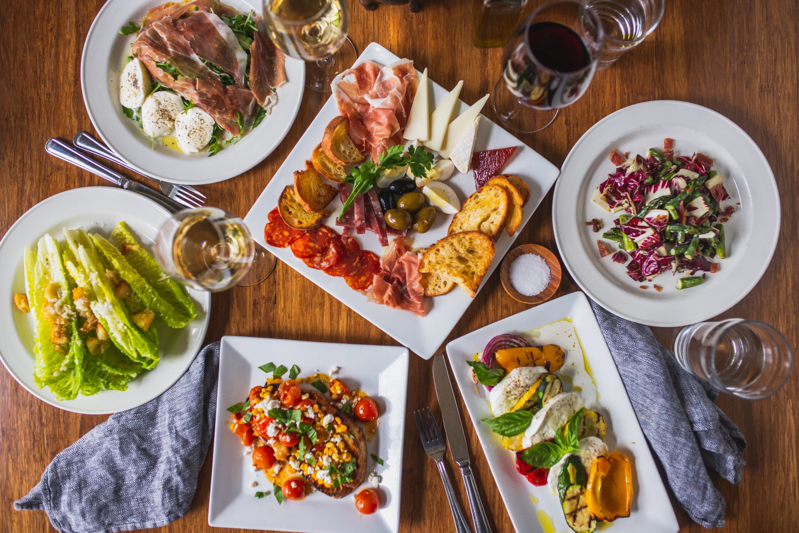 Assorted dishes including salad, charcuterie platter, and a bowl of soup on a wooden table with glasses of wine and utensils.