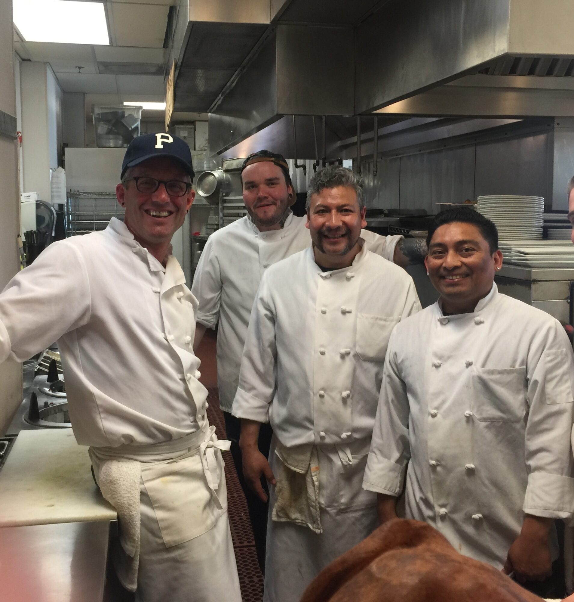 Four chefs in white uniforms standing in a commercial kitchen, smiling, with kitchen equipment and dishes in the background.