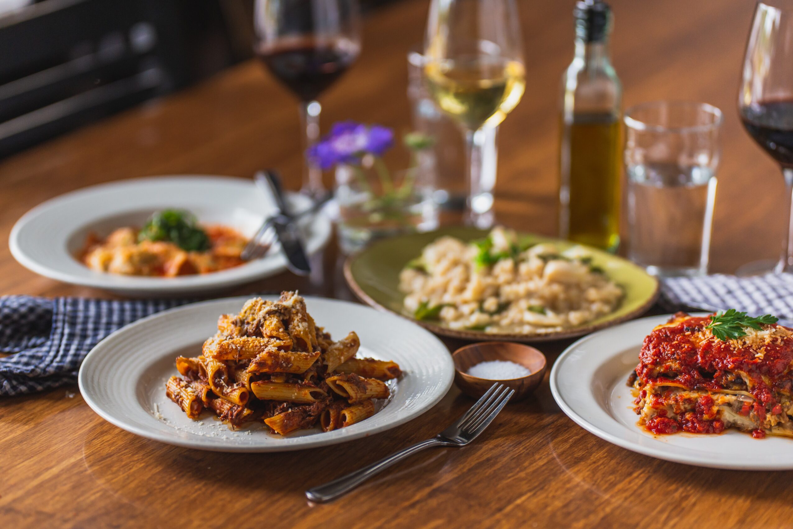 Three plates of food on a wooden table with wine glasses and bottles in the background.